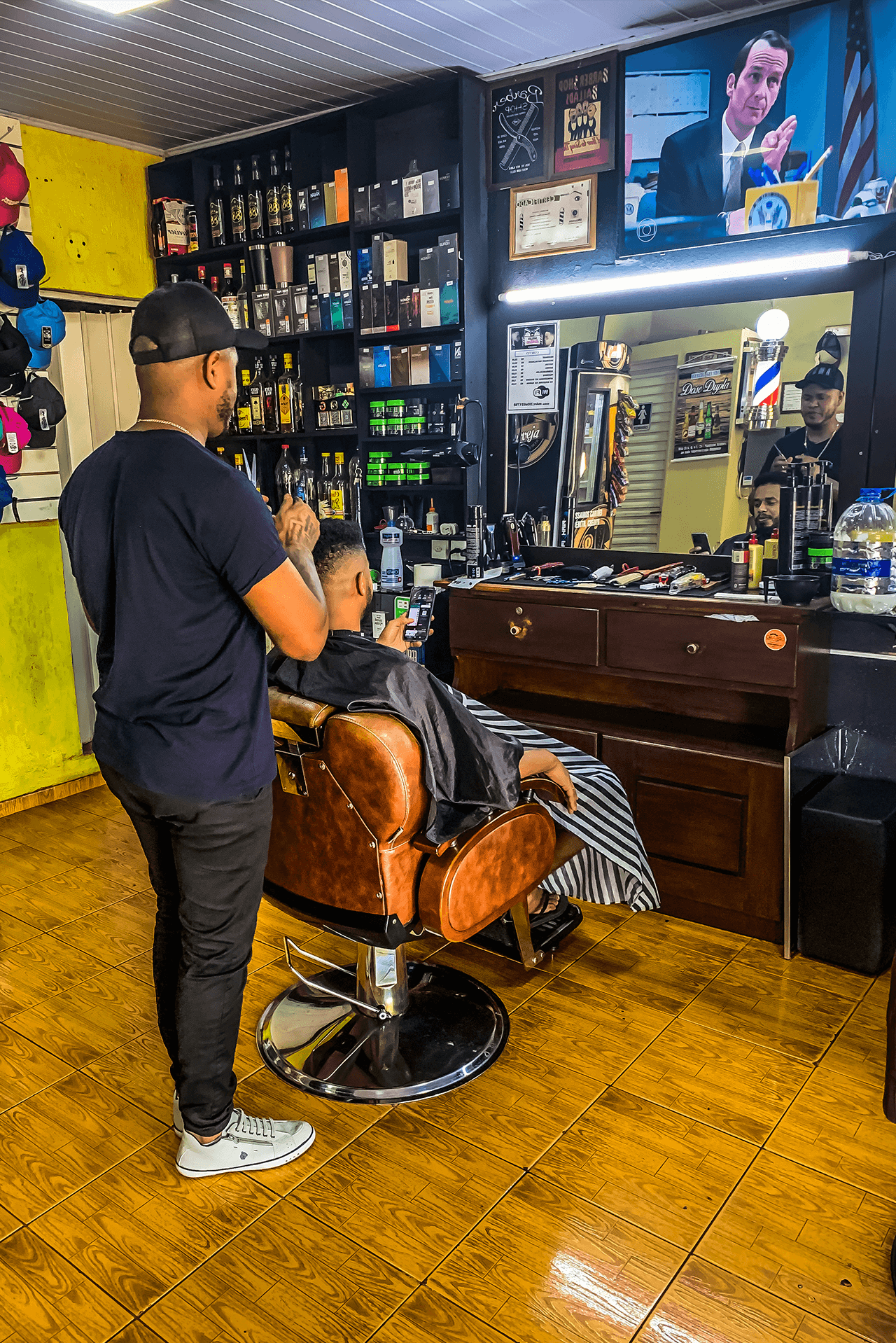 Marcelo, o experiente barbeiro e proprietário da Barbearia Santana em Rio Verde, realiza corte preciso no cabelo de um cliente acomodado confortavelmente na cadeira, enquanto acompanha um programa na televisão.